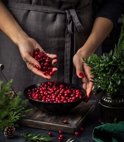 Ręce kobiety w szarym fartuchu delikatnie przesypują czerwone owoce żurawiny do czarnej miski na drewnianej desce, otoczonej świąteczną zielenią i szyszkami.