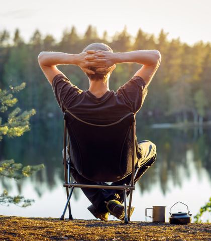 Mężczyzna odpoczywający na składanym krześle przy jeziorze, z rękami założonymi za głową, wpatrujący się w piękny, leśny krajobraz w popołudniowym świetle, co sugeruje spokojny czas na łonie natury.